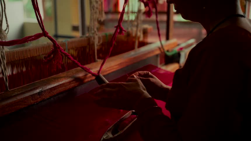 An artisan's hands carefully thread a traditional handloom in preparation for weaving a vibrant red fabric