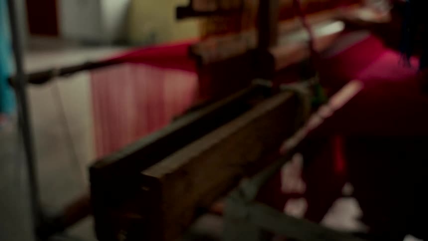A person skillfully operates a traditional wooden handloom, weaving vibrant red fabric in a workshop