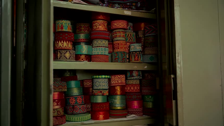 Rolls of ornate and colorful textile ribbons are neatly stacked on shelves in a storage cabinet