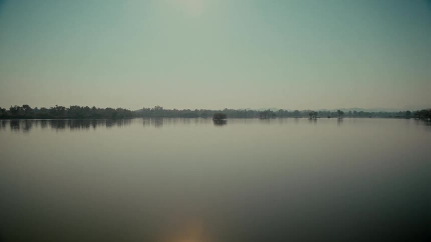 A wide, tranquil view shows a vast, calm lake with glassy water reflecting the hazy sky and distant shore