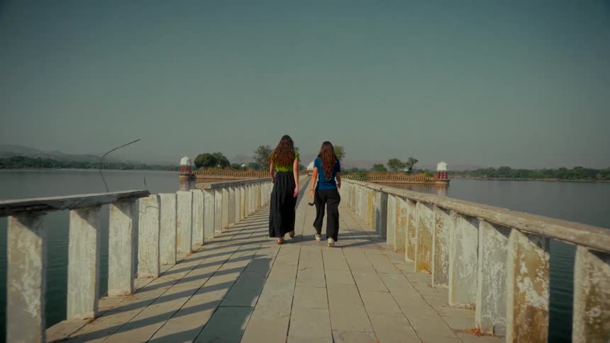 A slow-motion tracking shot from behind follows two women with long hair as they walk together down a historic stone causeway over a tranquil lake