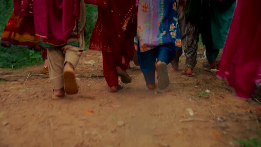 A low-angle shot from behind shows the feet of a group of women in colorful traditional clothing walking along a rural dirt path