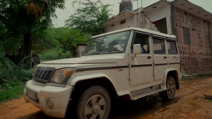 An SUV slowly drives past rundown buildings on a dirt road in a rural hillside village