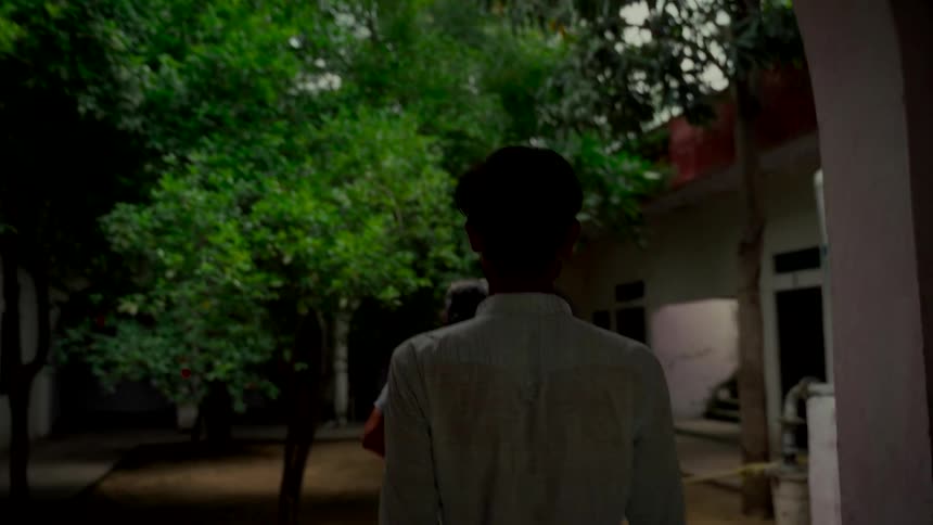 A young Indian man, seen from behind, walks in slow motion through an arched doorway into a lush, green courtyard