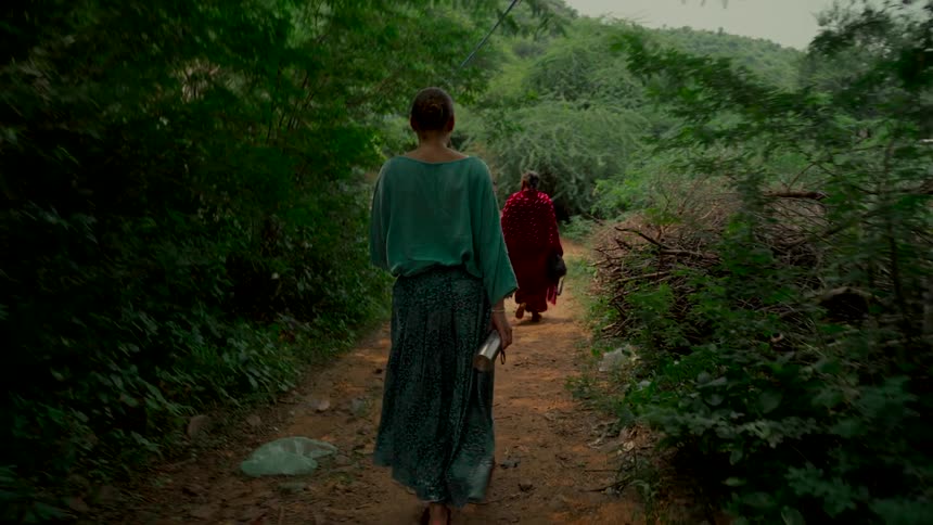 A woman in bohemian-style clothing follows a group of people down a dirt path through a lush green forest