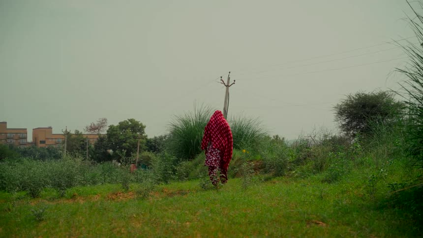 A person in a red traditional Indian garment walks through a green, rural landscape on an overcast day