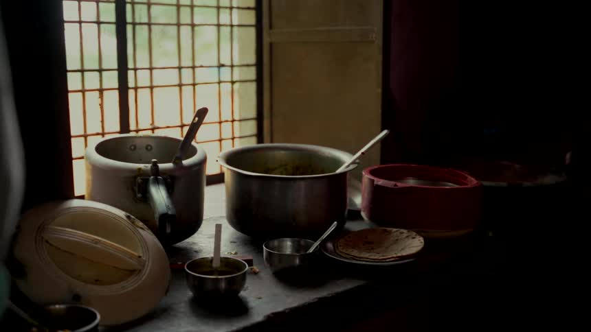 A still life of pots with food and fresh roti sits on a dark kitchen counter by a barred window as a fire burns in the background