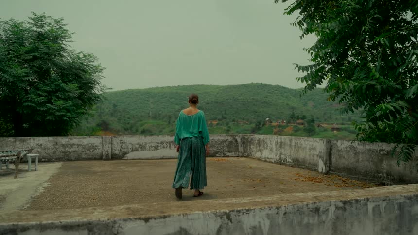 From behind, a woman in a flowing green outfit slowly walks across a rustic rooftop, admiring the view of the lush green hills