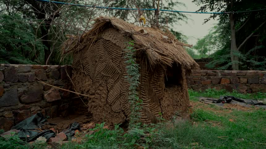 A traditional mud hut with a thatched roof and intricate tribal-style carvings sits in a rural, grassy area next to a stone wall