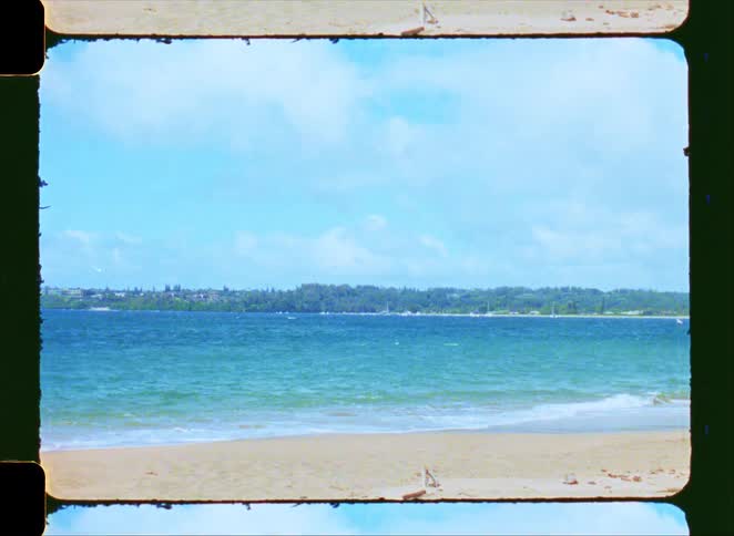 A serene, vintage film scan of a sandy beach with gentle waves lapping at the shore and a distant, tree-lined coast