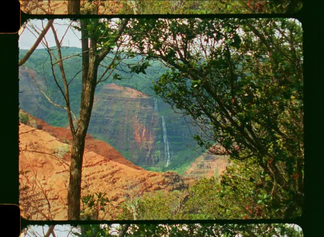 Vintage film footage slowly zooms out from a breathtaking waterfall flowing down a vibrant canyon wall, seen through a natural frame of trees