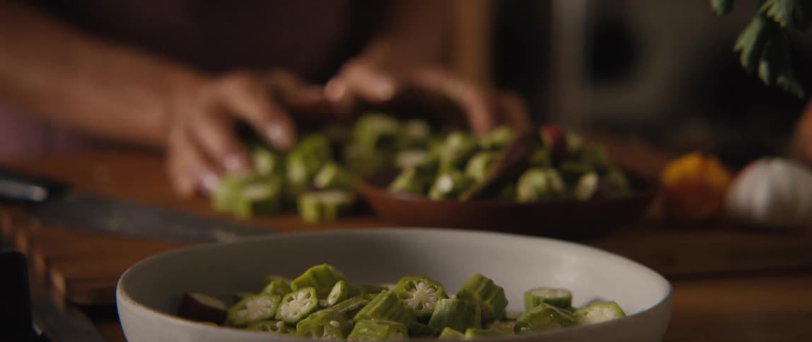 A close-up of a chef's hands chopping fresh green okra on a wooden cutting board and mixing it in a bowl