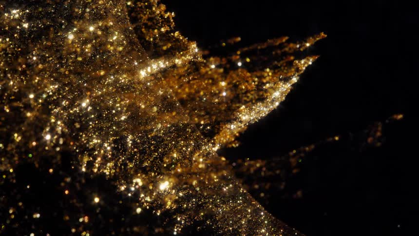 A cloud of shimmering gold particles and glitter floats gracefully in an abstract, slow-motion wave against a black background