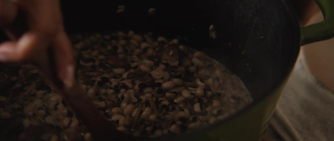 A woman lifts the lid of a green dutch oven to reveal a simmering pot of black-eyed peas, then stirs the stew with a wooden spoon