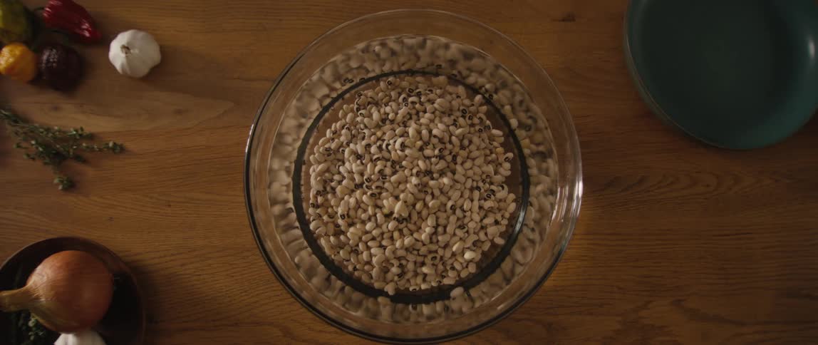 A top-down slow-motion shot of a person's hand washing black-eyed peas in a glass bowl of water on a wooden table