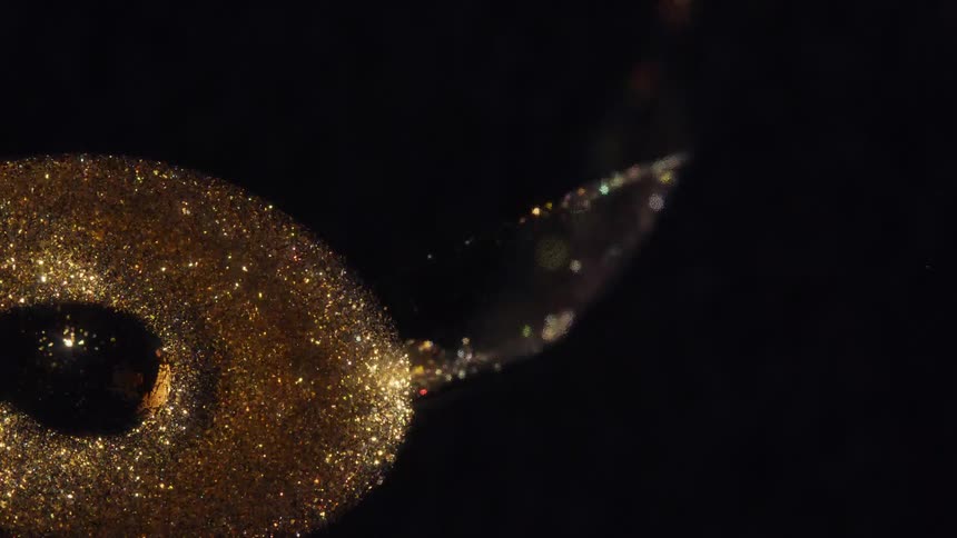 An artistic macro shot shows a brush smearing a thick, sparkling gold glitter paste across a black surface
