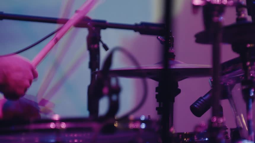 A drummer's hands move with speed and precision, playing a fast beat on the drum kit under vibrant purple stage lights