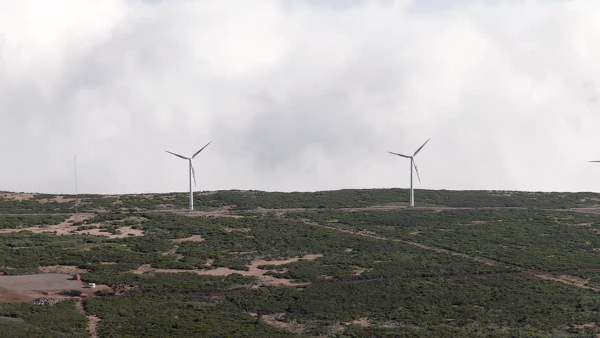 Wind turbines spin on a green hillside under a blanket of thick white clouds