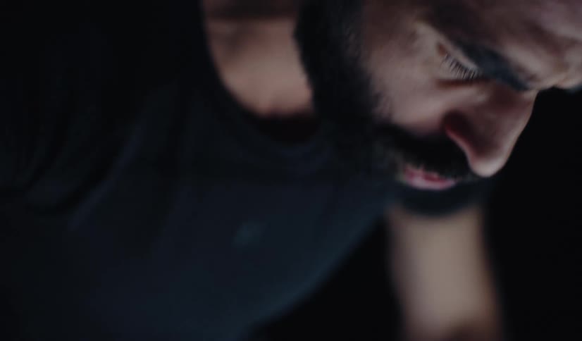 A shaky, high-angle close-up captures a bearded man's intense concentration in a dark room