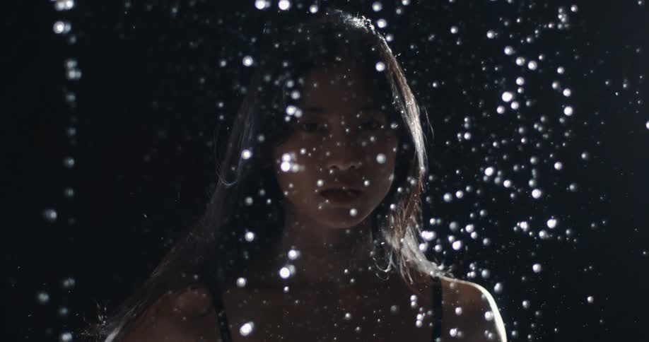 Cinemagraph portrait of a woman looking at the camera as illuminated water droplets fall in slow motion against a dark background