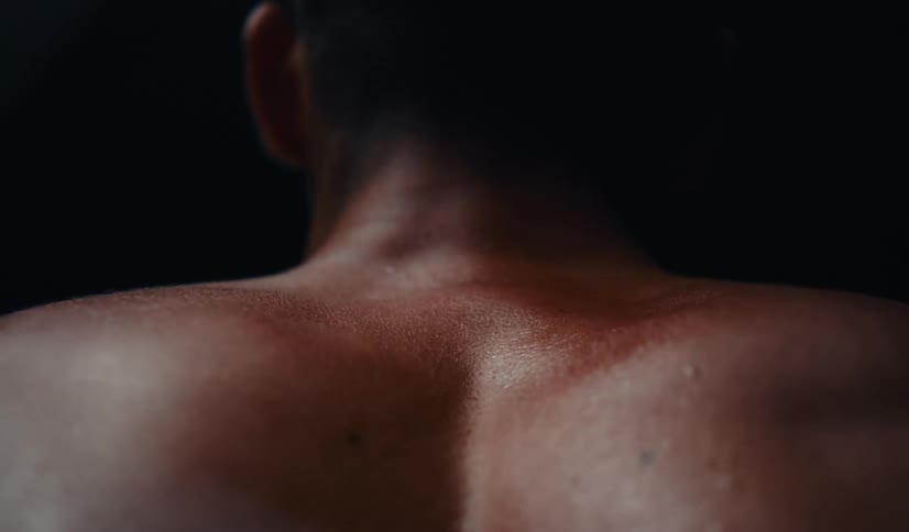 An artistic close-up from behind shows a man's sweaty back and shoulder muscles flexing during an intense workout