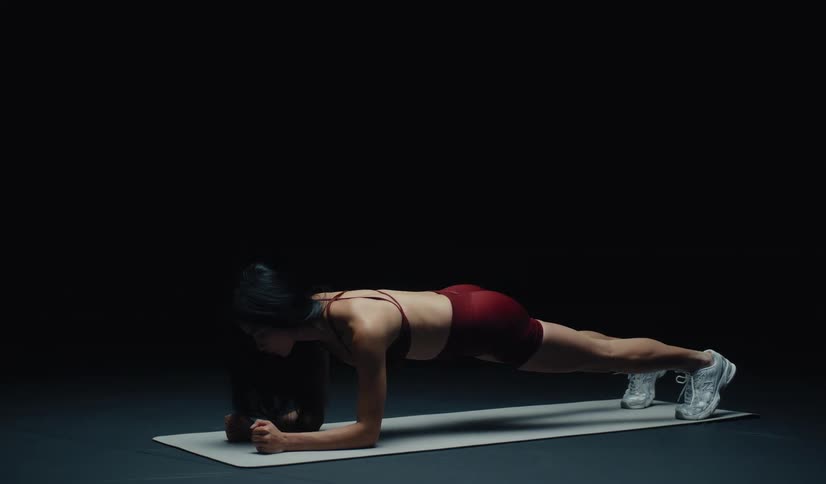 A determined woman in athletic wear performs plank up-downs on a yoga mat in a dark studio setting