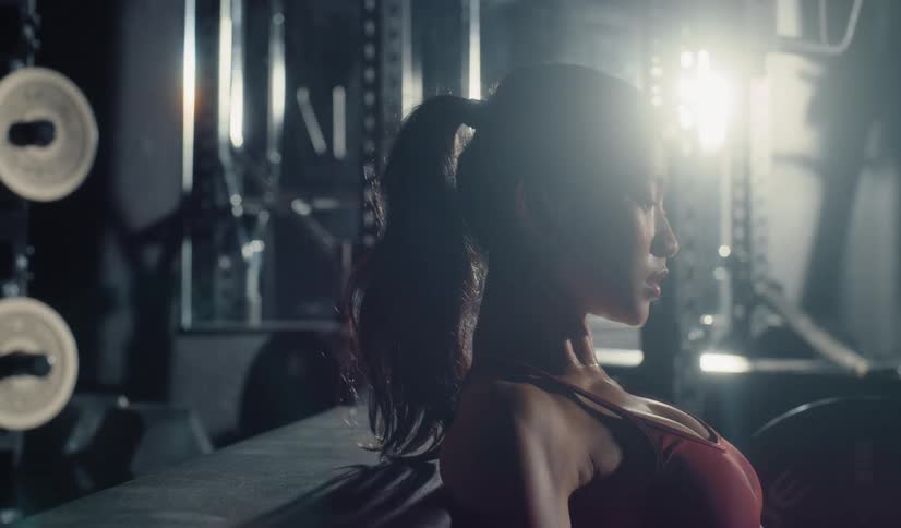 An athletic woman performs a set of intense hip thrusts in slow motion before taking a moment to rest in a dark, modern gym