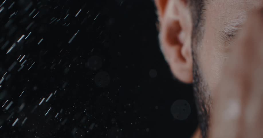 A dramatic slow-motion extreme close-up of a bearded man's intense face as rain falls on him against a black background