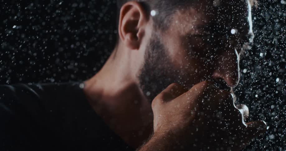 Super slow motion close-up of a determined, bearded man in a boxing stance getting drenched by rain against a black background
