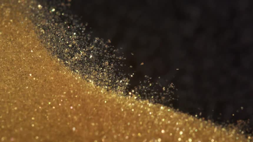 Mesmerizing macro slow-motion shot of sparkling gold glitter particles blowing away against a dark background