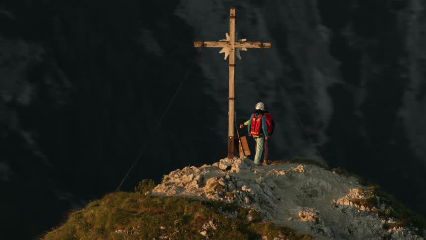 Two climbers stand next to a cross on a mountain peak
