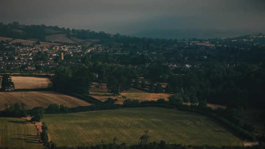 A slow aerial shot moves over the idyllic English countryside with a charming village nestled among the rolling hills and fields under a dramatic, overcast sky
