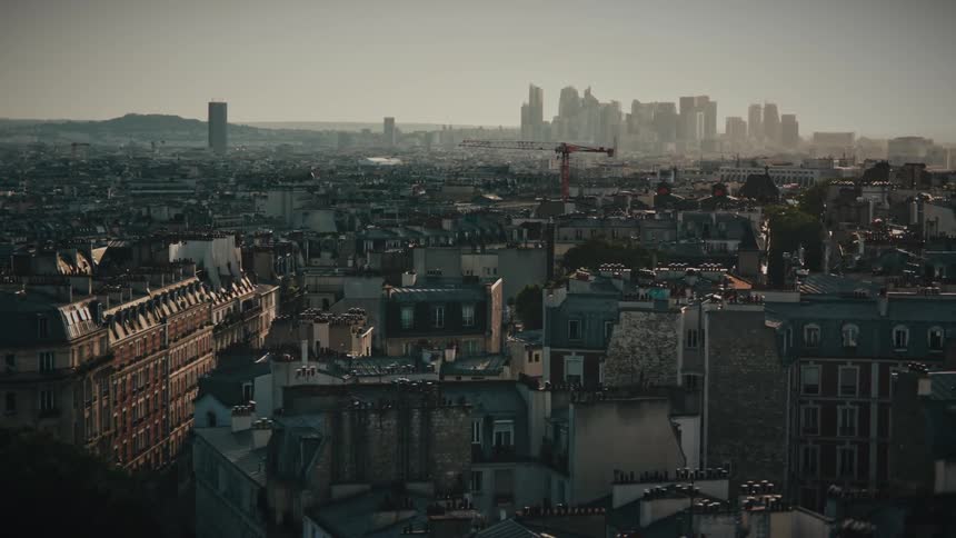 A slow aerial pullback over the classic rooftops of Paris reveals the modern La Défense business district on the horizon during a hazy golden hour