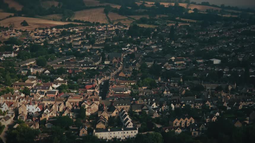 A high-angle, slow zoom-out shot reveals a quaint English town nestled among rolling green hills and fields