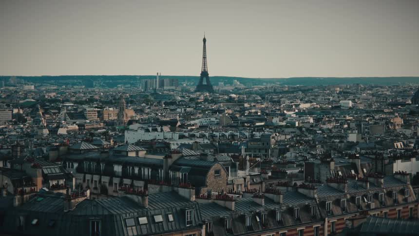 A slow aerial push-in shot over the rooftops of Paris, France, with the iconic Eiffel Tower visible on the horizon during a hazy day