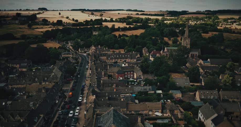 A slow aerial pan from left to right over a historic English village, revealing charming stone houses and a prominent church spire amidst the rolling green countryside