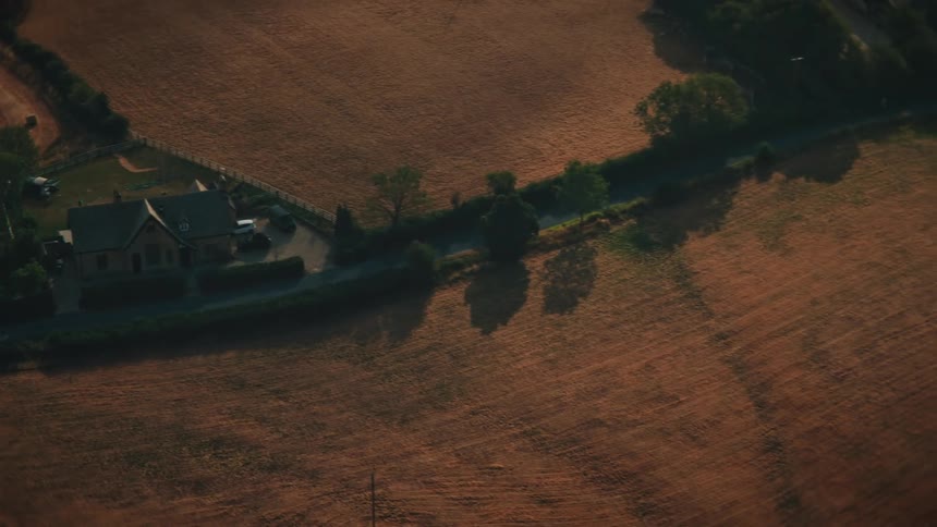 An aerial view of a harvested field with hay bales and a lone tree at sunset, pulling back to reveal a farmhouse and the vast rural countryside