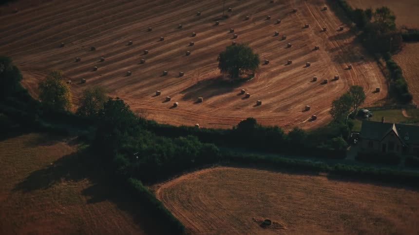 A slow aerial zoom captures the serene beauty of a rural landscape at sunset, focusing on a lone tree in a harvested field full of hay bales