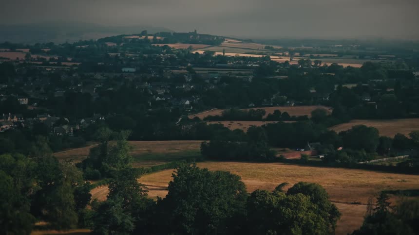 An aerial view pulling back from a quaint English countryside town nestled among rolling hills, fields, and a lush green park at dusk