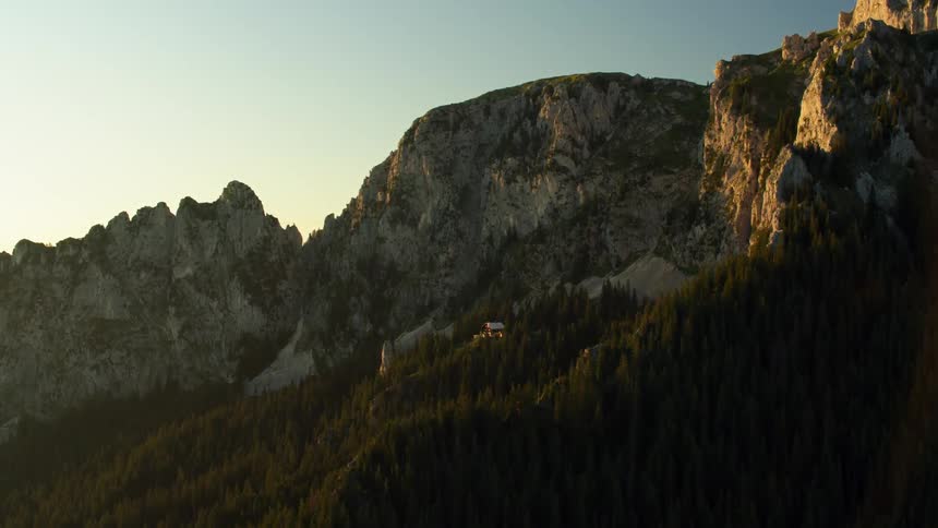 A cabin sits on a tree-lined cliffside below mountain peaks