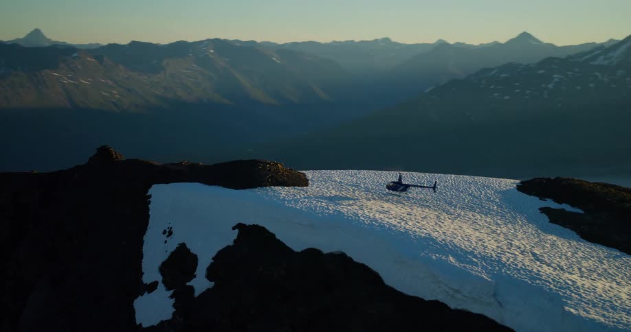 A helicopter rests on a snowy mountain peak at sunrise as the camera reveals a vast mountain range in the background