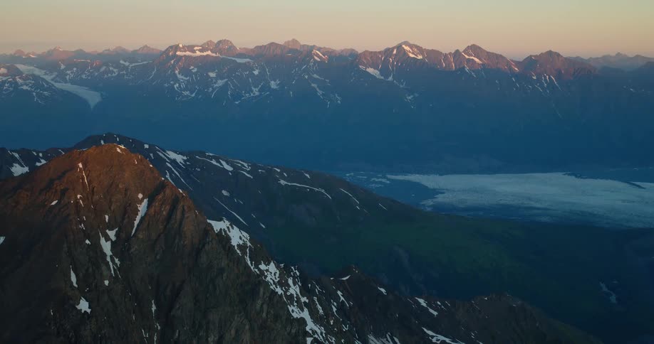 A breathtaking static shot of a vast, snow-capped mountain range at sunrise, with golden light illuminating the peaks and a glacier nestled in the valley below