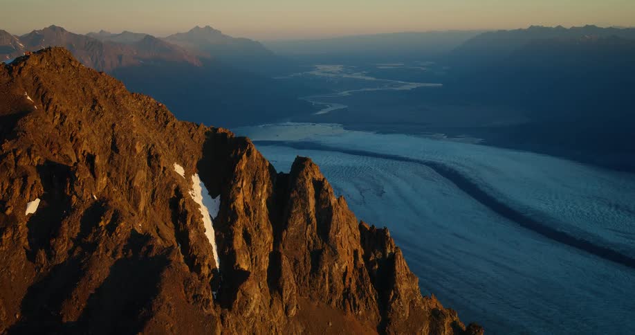 Golden hour light illuminates a craggy mountain peak, revealing a breathtaking view of a massive blue glacier flowing through a remote valley