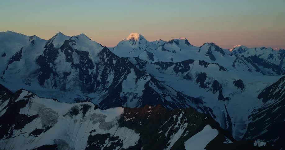 A serene wide shot of the last golden light of sunset hitting a distant snow-covered peak in a majestic mountain range