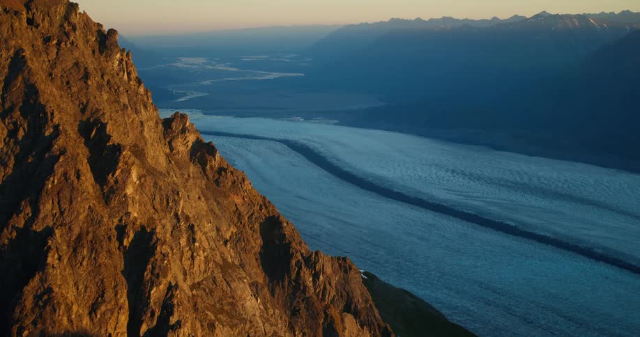 A stunning aerial view captures the golden light of sunset on a rugged mountain peak overlooking a vast blue glacier winding through a valley