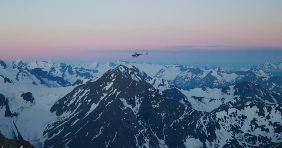 A helicopter soars over a majestic, snow-covered mountain range during a beautiful pink and blue sunset