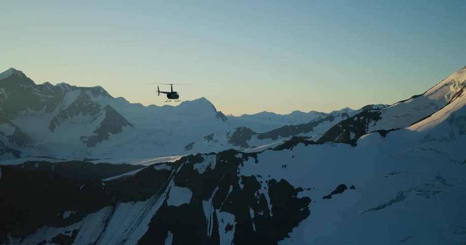 An aerial view of a helicopter flying over a vast snow-covered mountain range at sunset
