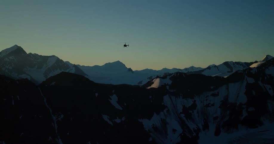 A silhouetted helicopter flies over a stunning, snow-covered mountain range at twilight