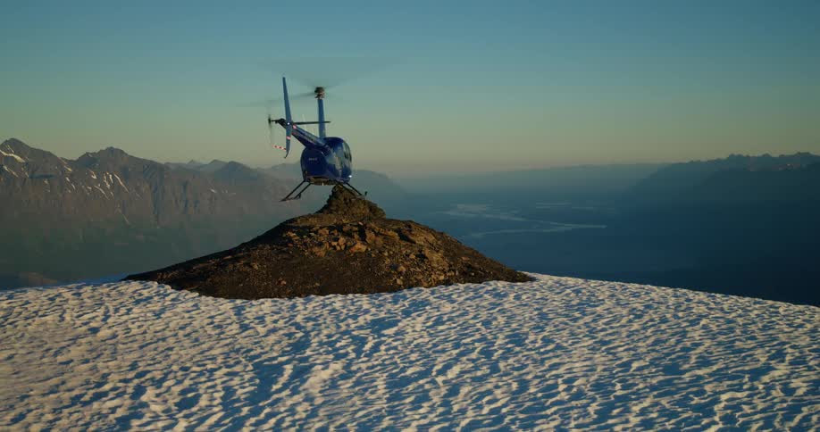 A blue helicopter takes off from a rocky peak on a snow-covered mountain, ascending over a vast glacier valley at sunrise