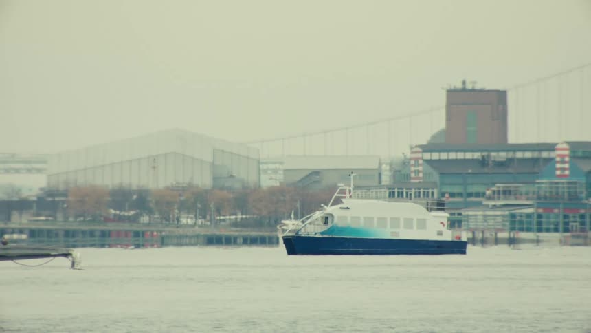 A slow-motion view of a commuter ferry crossing the river on a hazy, snowy day with the city shoreline in the background